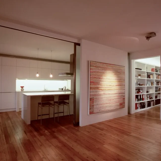 Interior de apartamento moderno con cocina minimalista, dos taburetes de bar en una isla, suelos de madera, obras de arte contemporáneo en la pared y estanterías empotradas en un pasillo bien iluminado.