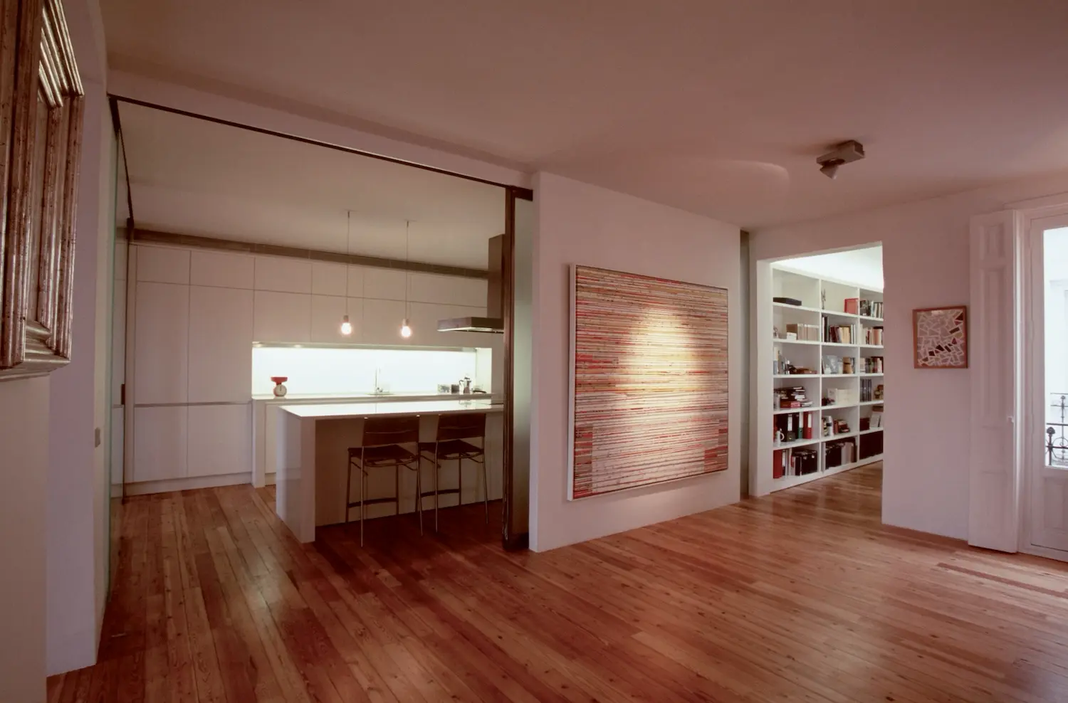 Interior de apartamento moderno con cocina minimalista, dos taburetes de bar en una isla, suelos de madera, obras de arte contemporáneo en la pared y estanterías empotradas en un pasillo bien iluminado.