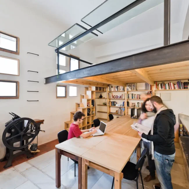 Un espacio de trabajo moderno y luminoso con techos altos, estanterías abuhardilladas, escaleras de madera y tres personas: una sentada en una mesa con un portátil y dos de pie mirando papeles. Los grandes ventanales dejan entrar la luz natural.