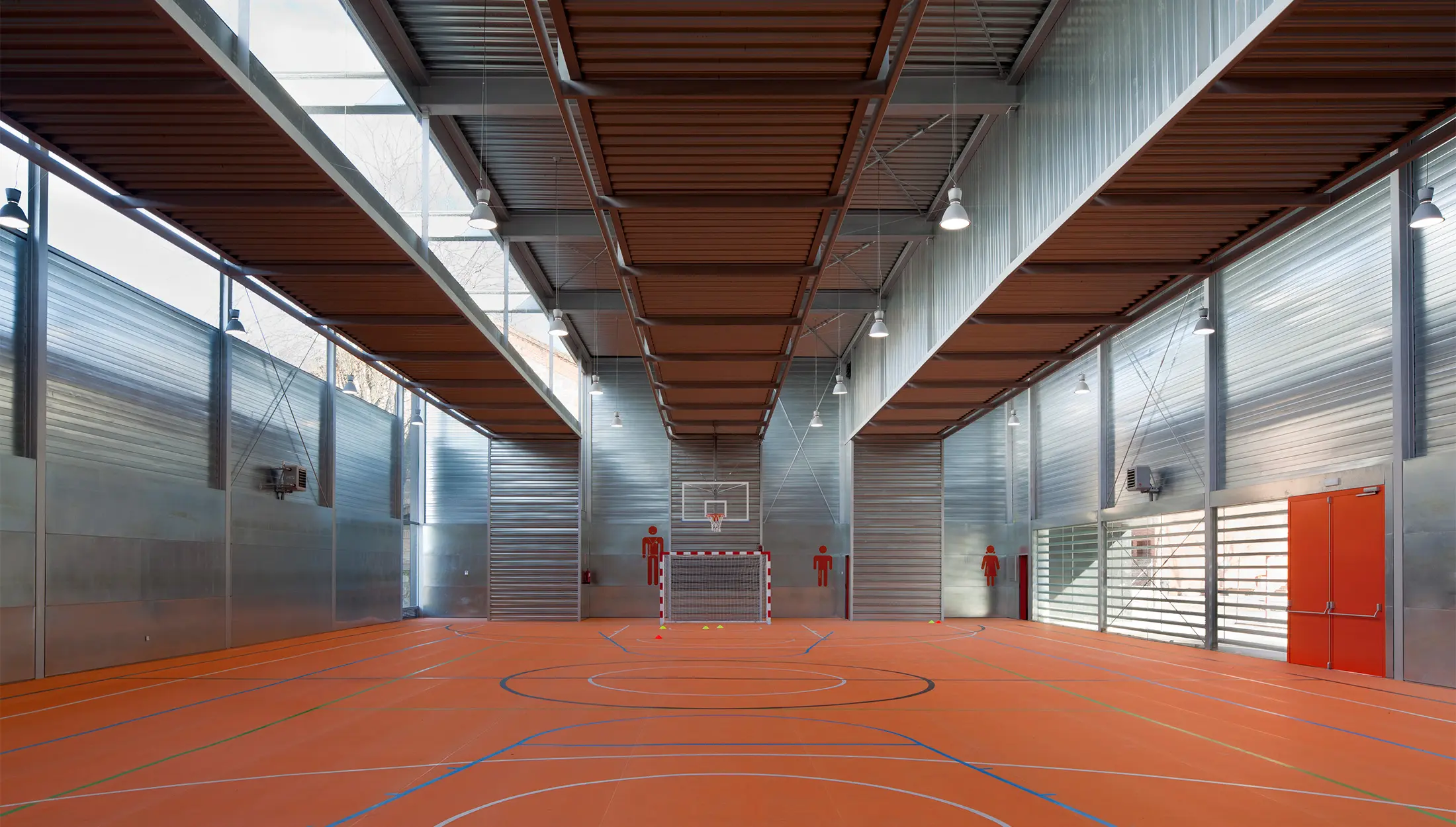 Un moderno gimnasio cubierto con suelo naranja marcado para varios deportes, altas paredes metálicas, grandes ventanales, luces cenitales y una canasta de baloncesto sobre un poste rojo y blanco en el fondo.