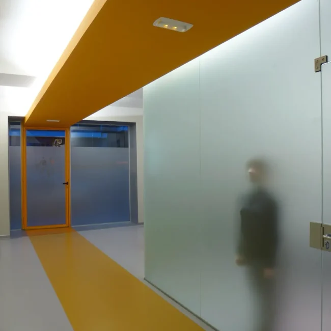Un pasillo moderno con el suelo y el techo amarillos y paredes de cristal esmerilado. Detrás del cristal esmerilado se ve una figura borrosa y, en primer plano, una puerta de cristal parcialmente abierta.