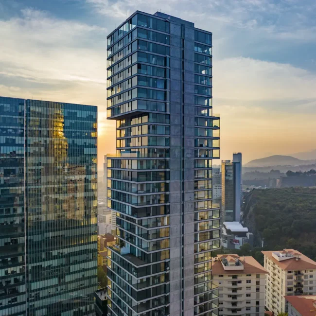 Altos rascacielos modernos de cristal con ventanas reflectantes al atardecer, rodeados de edificios residenciales más pequeños y vegetación, con montañas lejanas bajo un cielo parcialmente nublado.