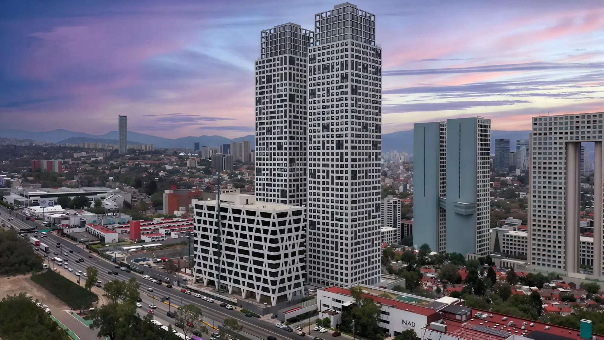 Dos rascacielos altos y modernos con un diseño cuadriculado se sitúan junto a un edificio más bajo de estilo similar. Los coches circulan por la carretera de enfrente, y el paisaje urbano se extiende hacia el fondo bajo un colorido cielo al atardecer.