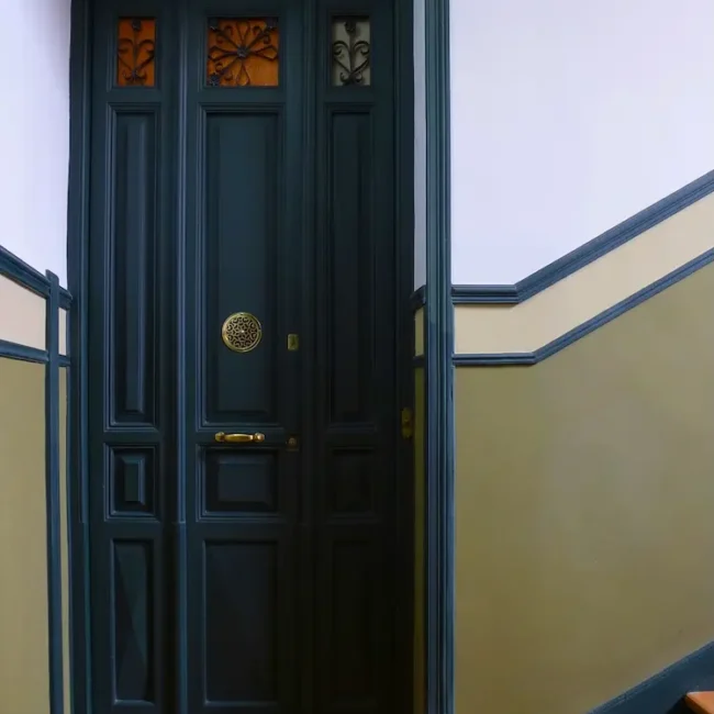 Una alta puerta de madera oscura con paneles dorados decorativos se encuentra en la parte superior de una escalera con paredes de color beige verdoso y suelo de madera. En la parte superior de la puerta hay pequeñas vidrieras.