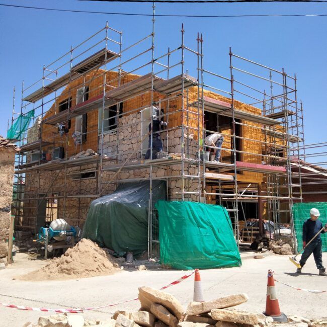 Una casa de piedra en construcción con andamios por todos lados. Hay trabajadores en los andamios y en el suelo. Hay montones de piedras, arena y materiales de construcción cerca, y conos de tráfico delimitan la zona. El cielo está despejado y azul.