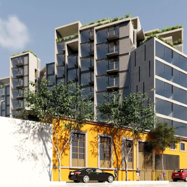 Un moderno edificio de varias plantas con balcones acristalados se alza detrás de una estructura amarilla de una sola planta. Hay coches aparcados a lo largo de la calle y árboles verdes bordean la acera bajo un cielo parcialmente nublado.