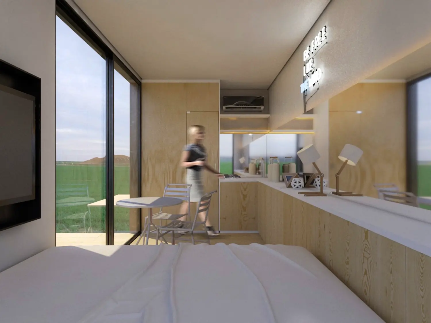 Una habitación de hotel moderna y minimalista con un gran ventanal que da a un campo cubierto de hierba. Una persona está de pie cerca de una cocina americana y una pequeña mesa con sillas. La cama está en primer plano y en la pared hay un televisor.