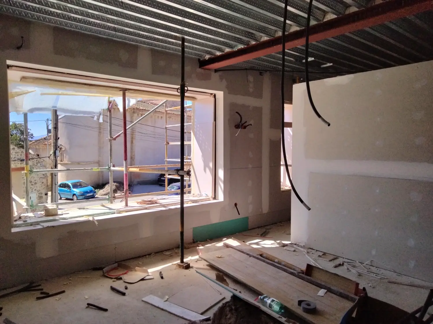 Una habitación en construcción con cables a la vista, paredes inacabadas y materiales de construcción esparcidos por el suelo. La luz del sol se cuela por una gran ventana, mostrando andamios y un coche azul aparcado fuera.