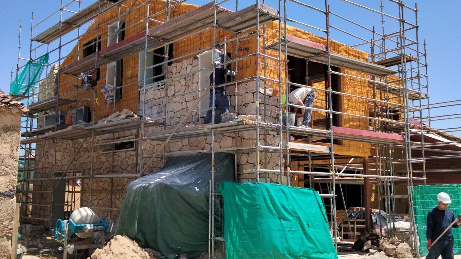 Una casa de piedra en construcción con andamios por todos lados. Hay trabajadores en los andamios y en el suelo. Hay montones de piedras, arena y materiales de construcción cerca, y conos de tráfico delimitan la zona. El cielo está despejado y azul.
