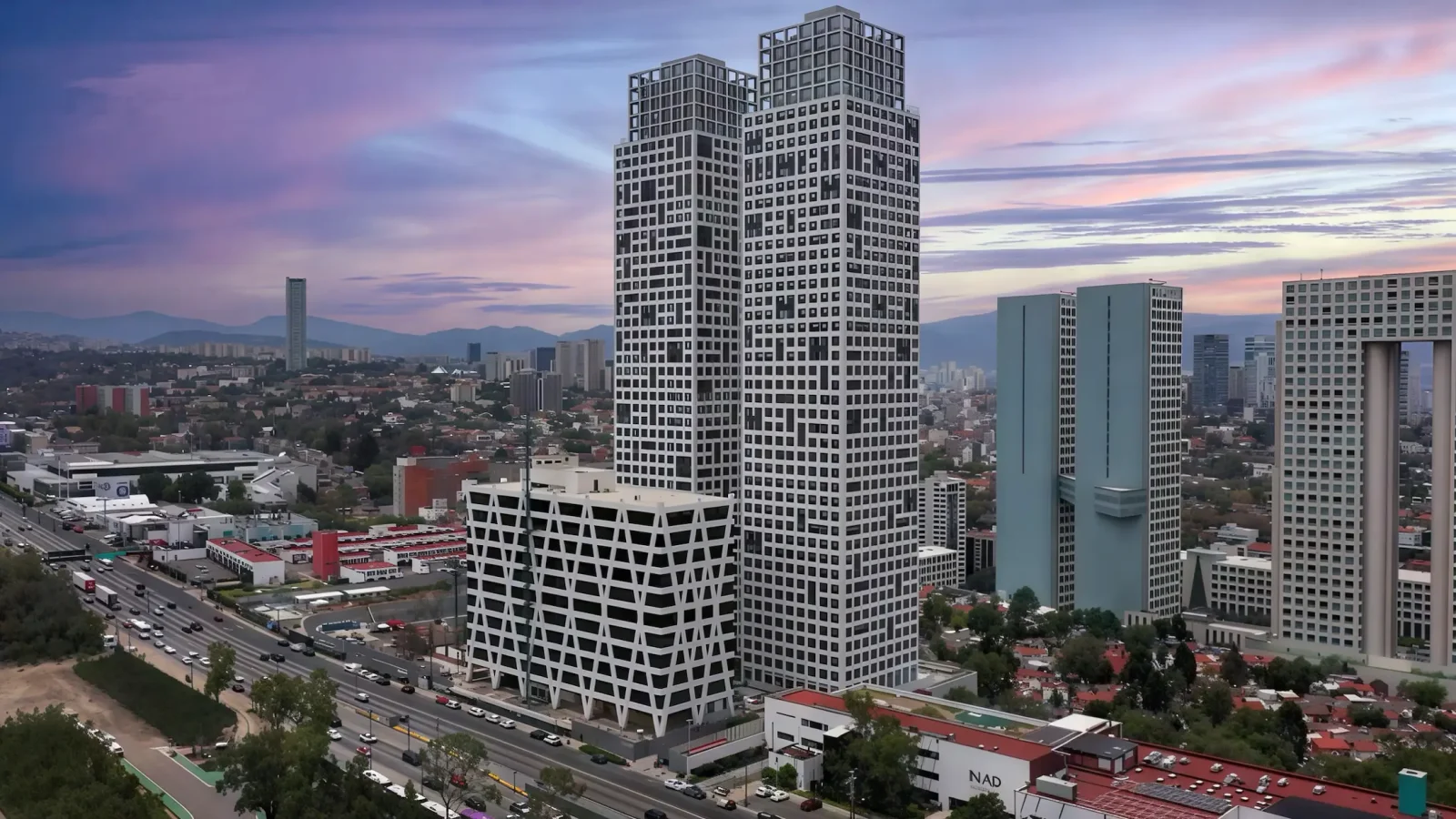 Dos torres de gran altura con ventanas geométricas junto a un edificio más bajo en un paisaje urbano al atardecer, con otros edificios modernos y montañas lejanas bajo un cielo colorido.