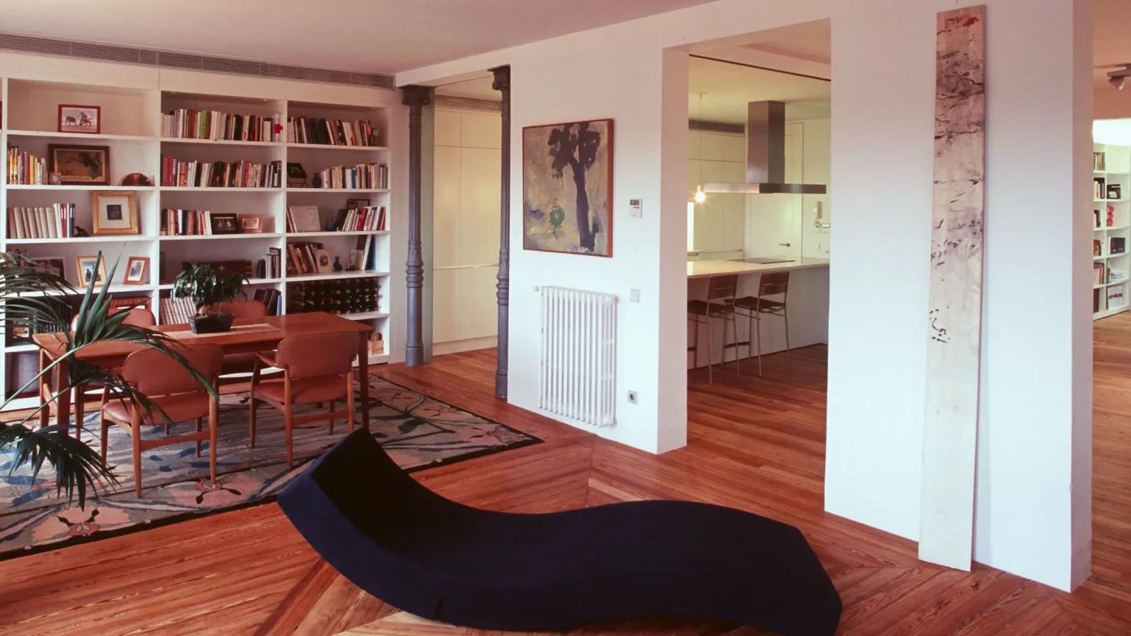 Un salón moderno con suelos de madera, una tumbona negra, estanterías, una mesa de comedor con sillas, obras de arte en las paredes y una cocina blanca a la vista.