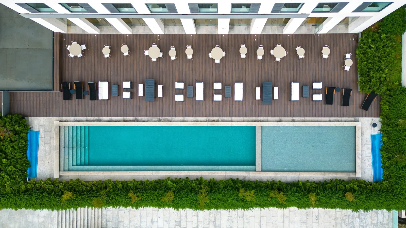 Vista aérea de una piscina rectangular rodeada de vegetación, con una cubierta de madera encima con mesas, sillas y sombrillas cuidadosamente dispuestas junto a un edificio blanco.