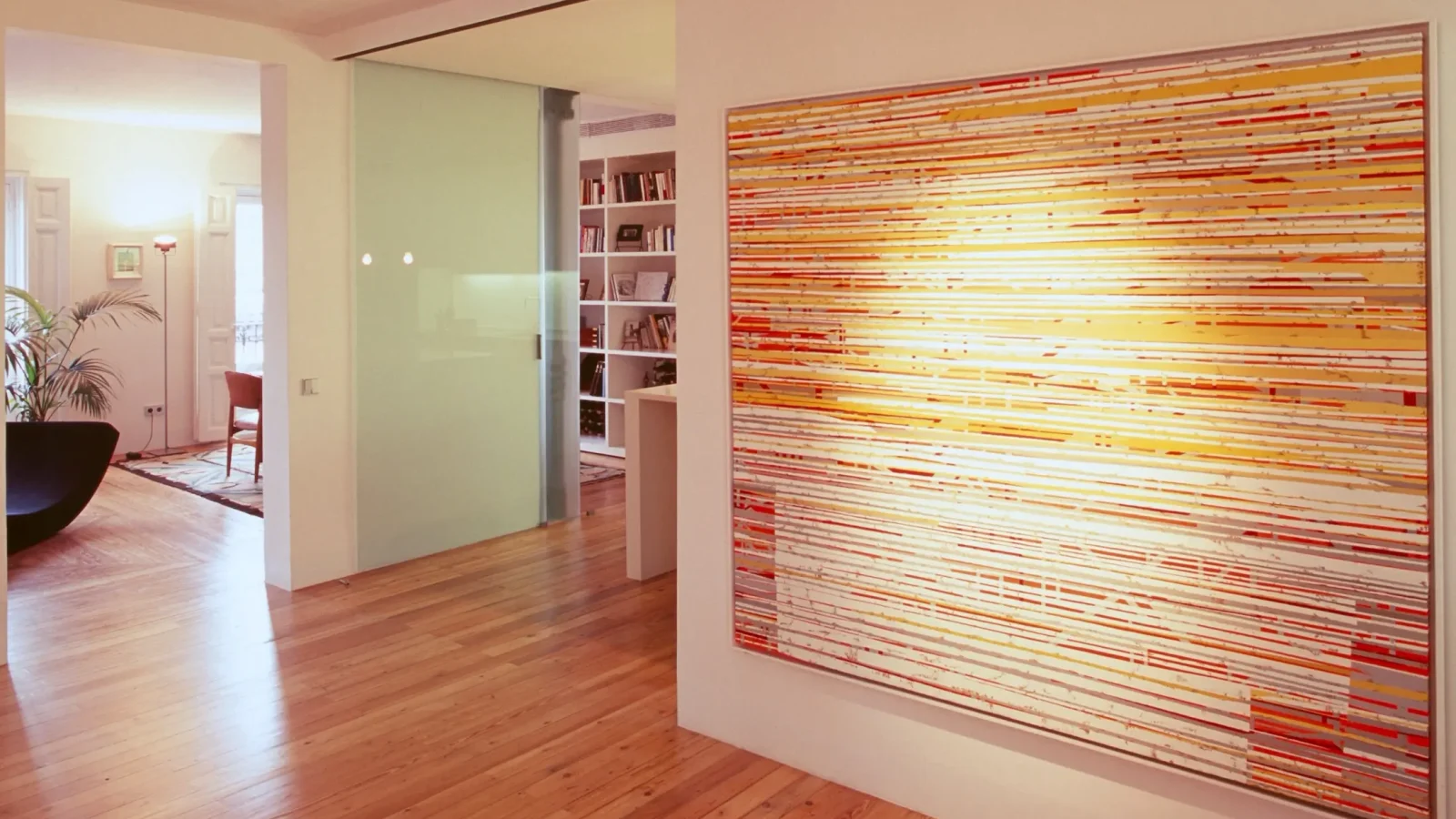Un salón moderno con suelos de madera, paredes blancas, un gran cuadro abstracto con líneas horizontales de colores, una puerta de cristal esmerilado y vistas a una habitación con estanterías y una silla negra.