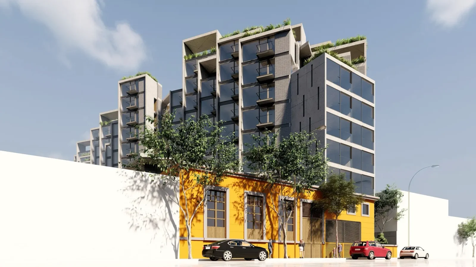 Un moderno edificio de varias plantas con balcones acristalados se alza detrás de una estructura amarilla de una sola planta. Hay coches aparcados a lo largo de la calle y árboles verdes bordean la acera bajo un cielo parcialmente nublado.