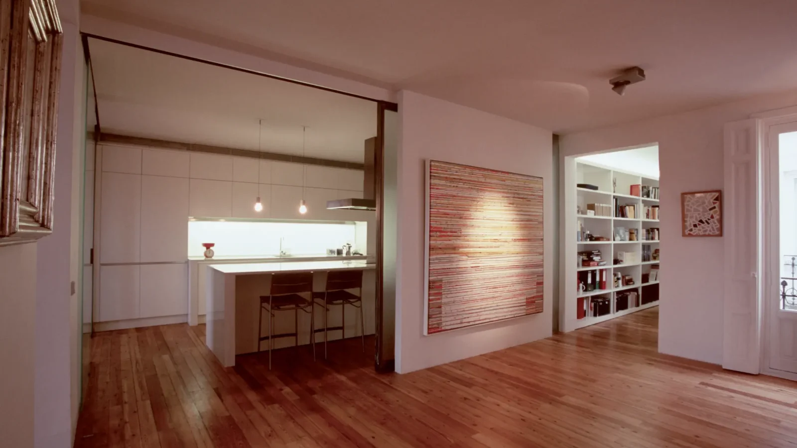 Moderno apartamento diáfano con suelos de madera, cocina blanca con isla y taburetes, lámparas colgantes, un gran cuadro abstracto de colores en la pared y estanterías empotradas en el pasillo.