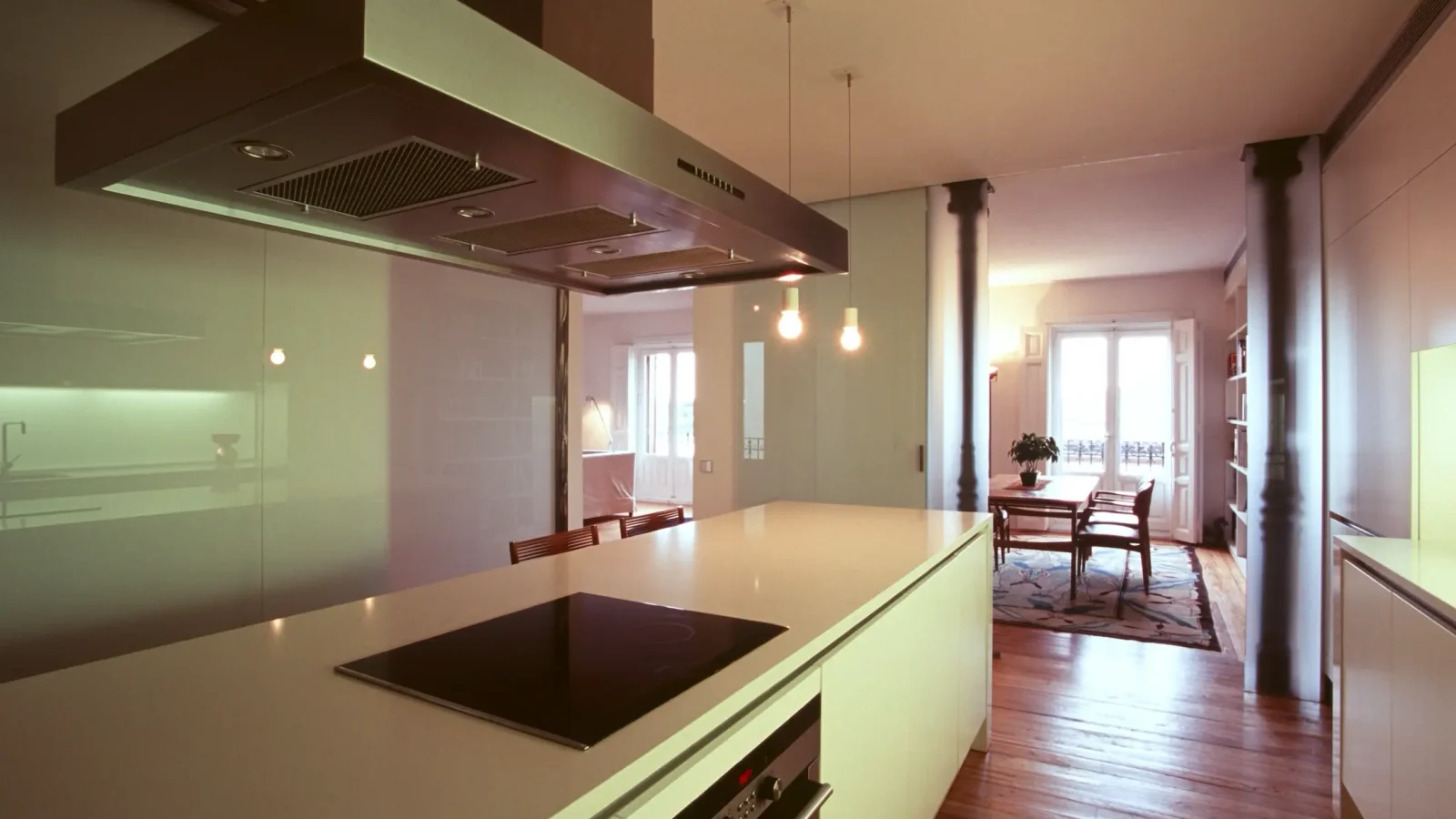 Cocina moderna con una elegante isla, placa de cocción integrada, campana extractora de acero inoxidable, lámparas colgantes y suelos de madera. Al fondo se ve un comedor abierto con grandes ventanales.