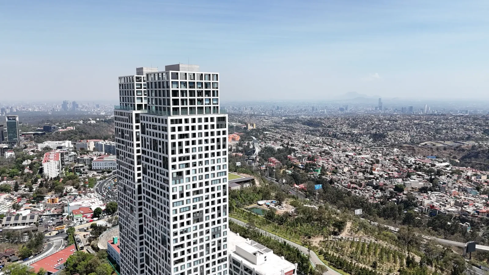 Un alto y moderno rascacielos blanco se alza en primer plano con un paisaje urbano en expansión y zonas verdes de parque visibles al fondo bajo un cielo azul despejado.