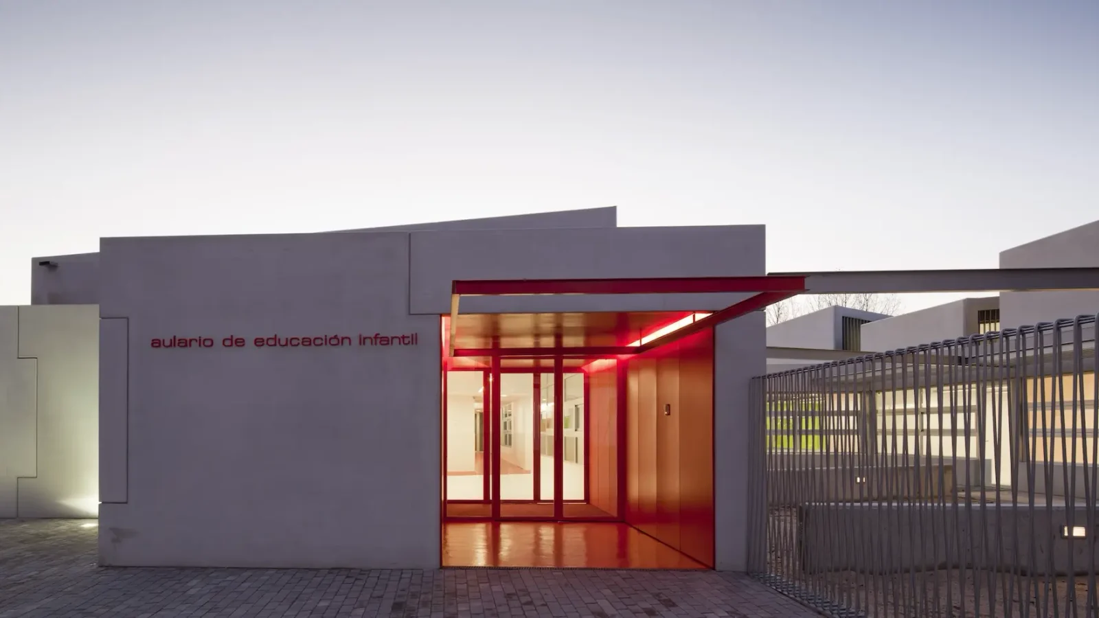 Un edificio moderno y minimalista con paredes blancas y una entrada con marcos rojos brilla con luz roja. El letrero dice "aulario de educación infantil", lo que indica que es un aula de educación infantil.