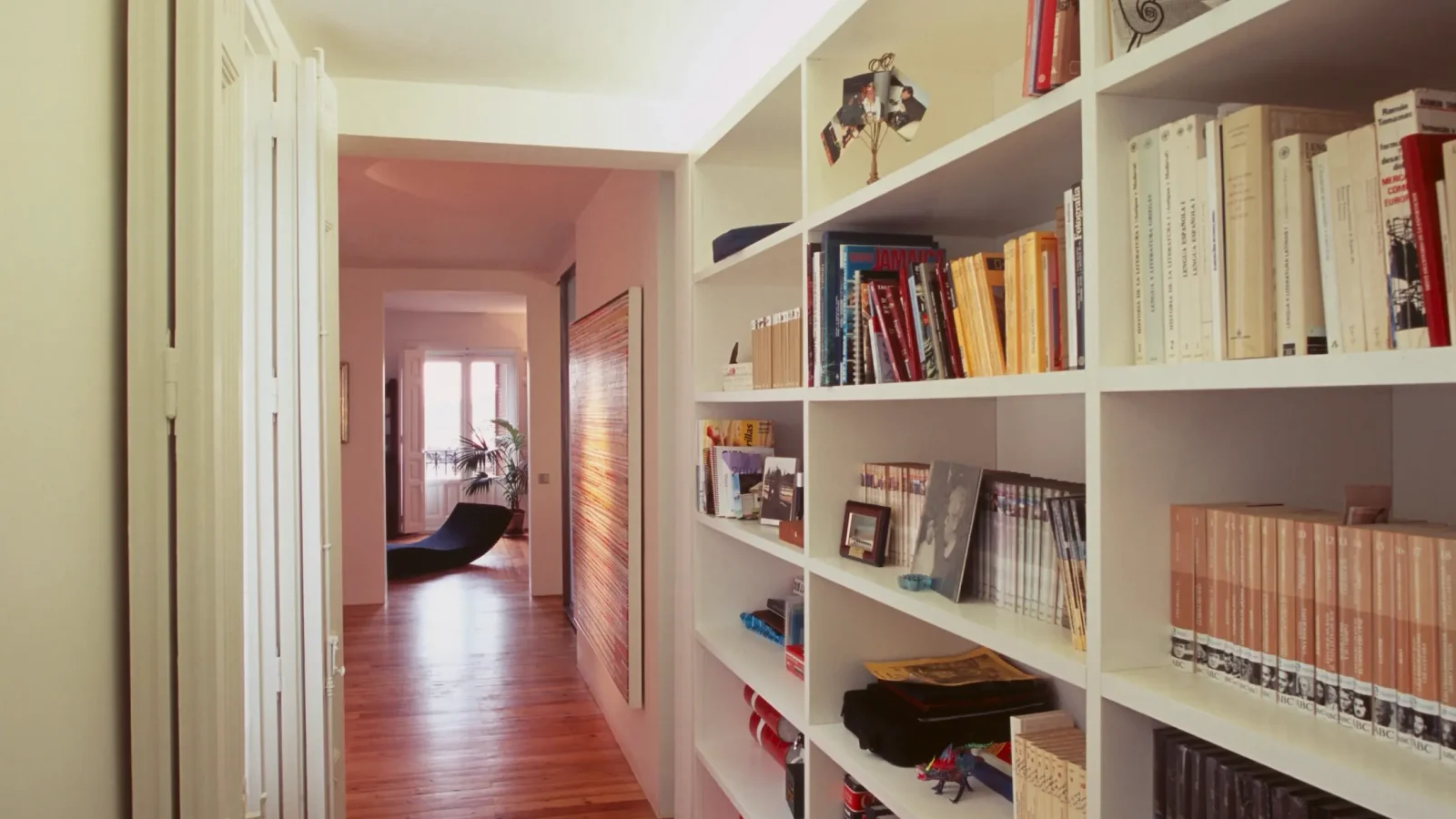 Un luminoso vestíbulo con suelos de madera y estanterías blancas empotradas llenas de libros y objetos diversos. El pasillo conduce a una habitación iluminada por el sol, con grandes ventanales y una silla negra.