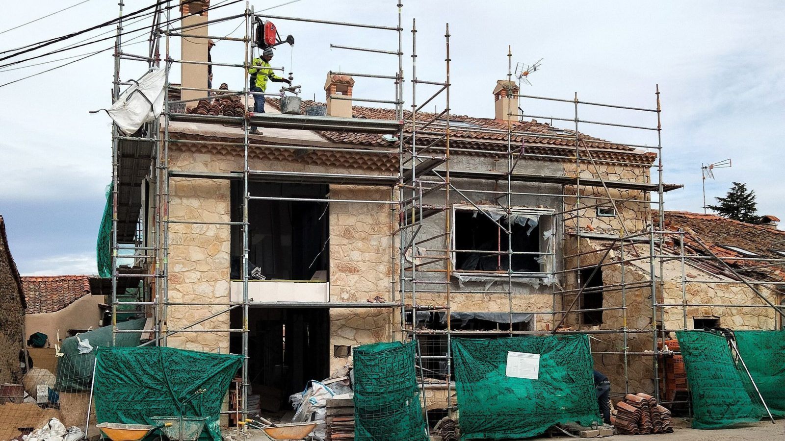 Una casa de piedra en obras está rodeada de andamios. Un obrero está en el tejado, y delante del edificio se ven materiales de construcción, carretillas y redes de seguridad verdes.