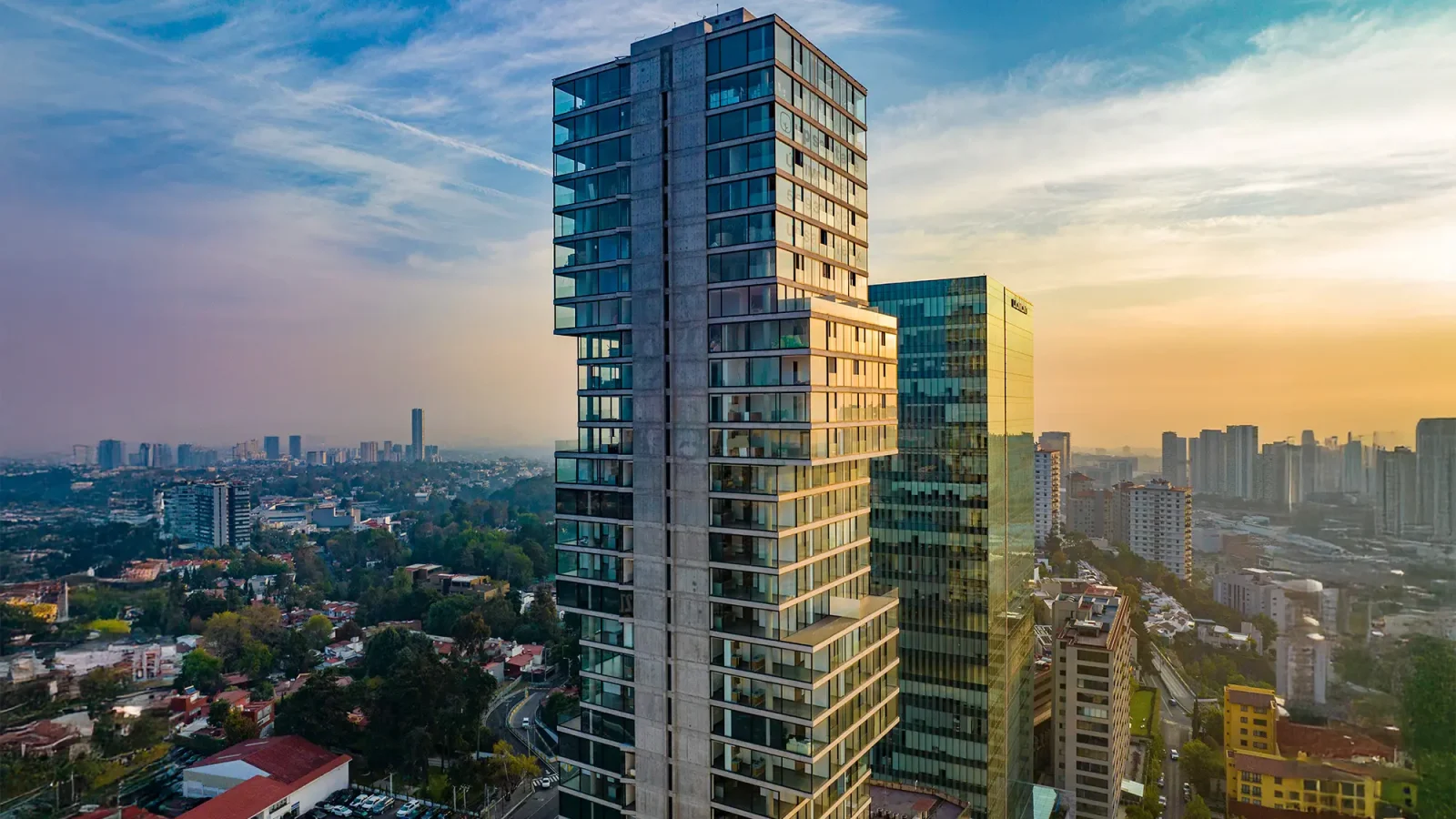 Modernos rascacielos con fachadas de cristal se alzan sobre un paisaje urbano al atardecer, rodeados de estructuras más bajas, vegetación y un horizonte lejano bajo un cielo azul y naranja.
